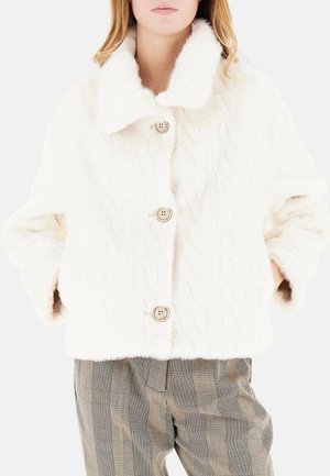 Giacca in pelliccia sintetica bianca con un ampio colletto, superficie texturizzata e chiusura con quattro bottoni. Indossata sopra pantaloni a quadri.