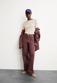 Wijde bordeauxrode jeans, geribbelde beige shirt, oversized bordeauxrode jas en een marineblauw pet met een glimlach-logo, gecombineerd met beige instap schoenen.