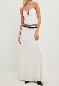 Top blanco sin tirantes con escote en forma de corazón, combinado con una falda maxi suave. Presenta un cinturón negro y unos tacones negros puntiagudos.