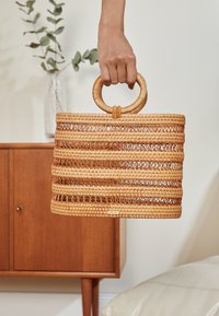 Håndtaske i flettet halm med cirkulært håndtag, der har vandrette striber og åbne fletninger. Naturlig tan-farve. Kompakt, struktureret form.