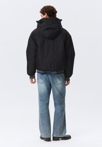 Sort isoleret jakke med hætte, der har elastiske manchetter og kort skæring. Sammen med lyseblå slidte jeans og sorte fodtøj.