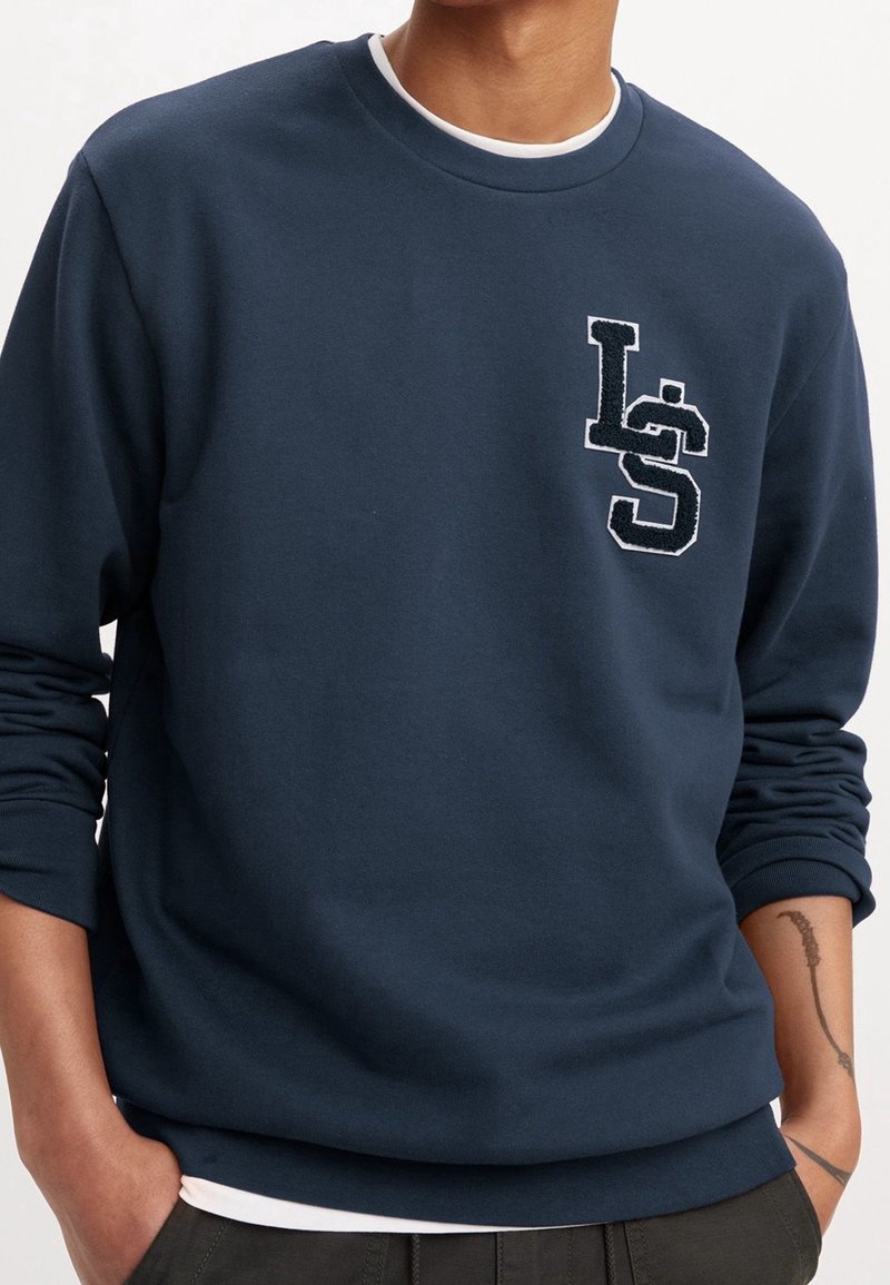Personne portant un sweat-shirt bleu marine avec les manches retroussées, avec un logo texturé « LS » sur la partie supérieure gauche de la poitrine.
