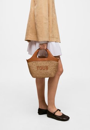 Mujer con chaqueta beige y falda blanca sosteniendo un bolso tote tejido con ribete de cuero y logo "TOUS", calzando zapatos Mary Jane negros.