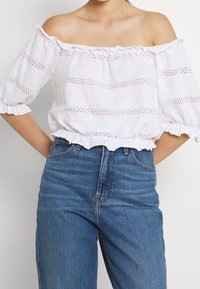 Bílá blůza s odhalenými rameny a volány, s očkovými vzory a přiléhavým pasem, spárovaná s modrými džínami s vysokým pasem.