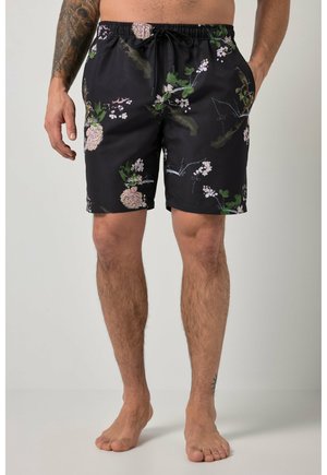 Hombre con pantalones de baño negros con estampado floral y cintura con cordón, de pie descalzo, mostrando su antebrazo izquierdo y tobillo tatuados.