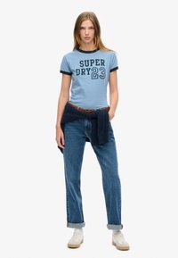 Lichtblauw t-shirt met korte mouwen en donkerblauwe rand met de opdruk "SUPER DRY 23". Draagt het met blauwe jeans en een marineblauwe trui om de taille geknoopt.