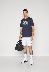 Marineblauwe t-shirt met wit PUMA-logo, gecombineerd met witte shorts. Zwarte sporttas en witte schoenen met rode accenten. Korte mouwen en zichtbare tattoos.