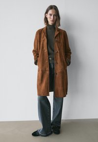 Bruine suède overjas met een losse pasvorm, voorzien van knopen, zijzakken en een gladde textuur, gecombineerd met een grijze geribbelde coltrui en wijde jeans.