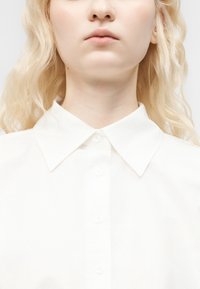 Ξανθό άτομο με ανοιχτά μαλλιά που φοράει ένα λευκό πουκάμισο με γιακά και κουμπιά, εμφανίζοντας τον λαιμό και το κάτω μέρος του προσώπου με ουδέτερη έκφραση.