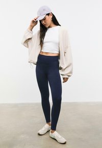 Lichtbeige cropped bomberjack, witte fitted top, marineblauwe leggings en grijze pet; heeft een relaxte pasvorm, zachte stof en casual sneakers.