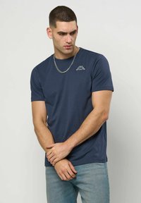 Νέος άνδρας που φοράει μπλε σκούρο t-shirt Kappa και ανοιχτό μπλε τζιν, στέκεται με τα χέρια σταυρωμένα, κοιτάζοντας προς τα κάτω δεξιά.