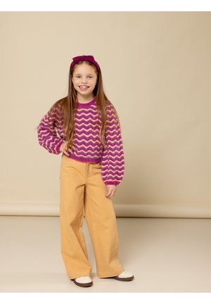 Meisje in een paarse geblokte trui, beige wijde broek, witte schoenen en een bijpassende paarse haarband, staand met een hand op de heup.