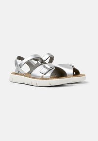 Silver metallic sandals with crisscross straps, a cutout design, and a white rubber sole. Features a cork insole and adjustable buckle.