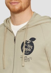 Sweat à capuche zippé khaki avec un grand graphisme bleu portant l'inscription "The Big Apple N.Y.C." sur la poitrine. Il dispose d'une capuche avec cordon de serrage et d'un tissu texturé.