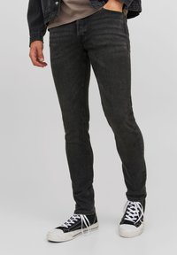 Blugi negri din denim, cu o croială slim, având o textură subtilă și detalii de cusături, asortați cu adidași negri și albi.