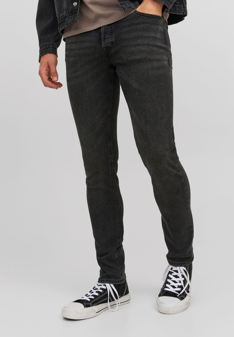 Blugi negri din denim, cu o croială slim, având o textură subtilă și detalii de cusături, asortați cu adidași negri și albi.