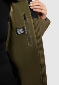 Olivengroene pufferjack met een gewatteerde textuur, voorzien van een ritszak en een merketiket op de linkerarm. Glad, glanzend materiaal.