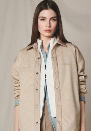 Kvinde med langt brunt hår iført lagdelte beige og blå lynlåstrøjer, stående mod en ensfarvet neutral baggrund og kigger fremad.