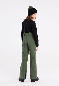 Pantalons de neige isolés verts avec bretelles ajustables et une forme évasée, accompagnés d'un bonnet tricoté noir et vert. Accents noirs au niveau des chevilles.