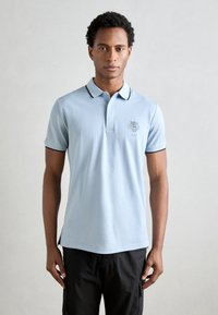 Polo de color azul claro hecho de algodón, con cuello, mangas cortas y un ribete negro. Incluye un logo bordado en el pecho.