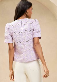 Lavendelbluse mit Puffärmeln, die mit gestickten Blumen- und Zickzackmustern, Ausschnitten und einer Schlüssellochöffnung am Rücken ausgestattet ist.