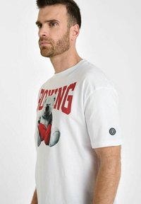 Camiseta blanca de algodón con un gran gráfico de un oso sosteniendo un corazón rojo, con la palabra "BOXING" impresa en letras rojas en negrita arriba. Mangas cortas.