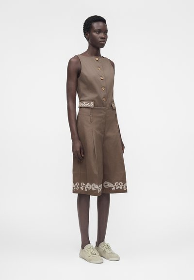 Γυναίκα που φοράει αμάνικο καφέ φόρεμα μέχρι τα γόνατα με κουμπιά στο μπροστινό μέρος, κεντητό τελείωμα και τσέπες, σε συνδυασμό με μπεζ αθλητικά παπούτσια, στέκεται ακίνητη.