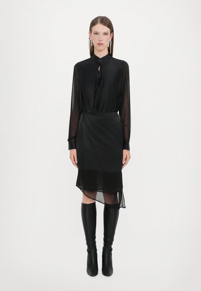 Blusa nera con collo legato, maniche lunghe trasparenti, gonna stratificata trasparente con orlo asimmetrico, abbinata a stivali neri alti.