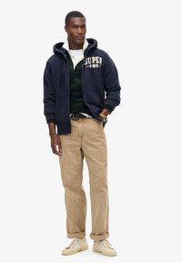 Felpa con cappuccio blu navy con logo, indossata sopra una maglietta verde scuro, abbinata a chinos beige e scarpe da ginnastica color crema, con i polsini arrotolati.