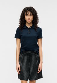 Polo bleu marine avec un col classique et trois boutons dorés, associé à un short noir à fines rayures avec une taille haute et des boutons sur le devant.