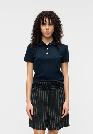 Marineblaues Poloshirt mit klassischem Kragen und drei goldenen Knöpfen, kombiniert mit schwarzen Shorts mit Nadelstreifen, hoher Taille und vorderen Knöpfen.