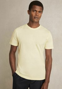 Cămașă galbenă cu mânecă scurtă și guler rotund, fabricată dintr-un material moale. Modelul pozează cu mâinile în buzunare, purtând pantaloni negri.