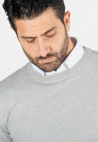 Pull en tricot gris avec un col rond côtelé, porté par-dessus une chemise blanche à col, présentant une texture lisse et une silhouette ajustée.