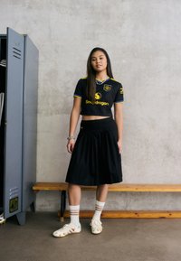 Jersey preto cortado com detalhes em amarelo e logotipo, combinado com uma saia plissada preta e sapatilhas de futebol em creme. Meias brancas com riscas castanhas.