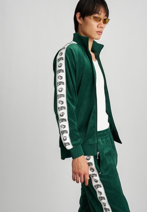 Grüner Samt-Trainingsanzug mit weißen Seitenstreifen und Logo. Die Jacke hat einen hohen Kragen und einen durchgehenden Reißverschluss; die Hose hat einen elastischen Bund.