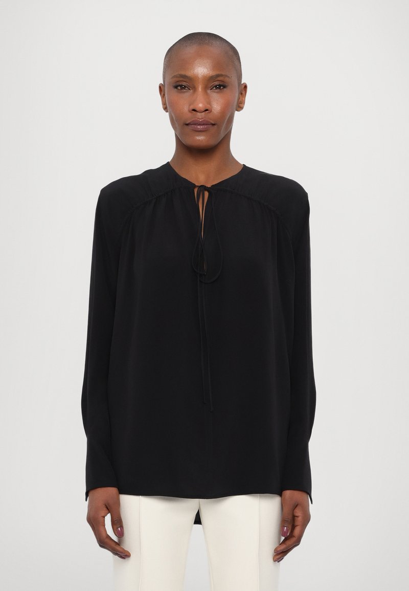Blusa nera a maniche lunghe con collo rotondo, caratterizzata da una chiusura con fiocco frontale e dettagli arricciati lungo le spalle. Tessuto setoso.