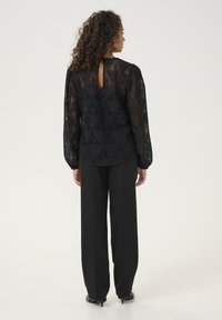 Haut en dentelle noire avec des manches longues, un col rond et un détail en forme de trou de serrure à l'arrière, associé à un pantalon noir ample.