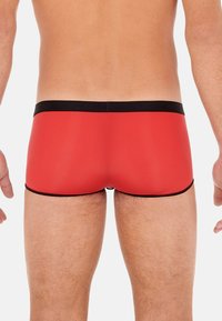 Red swim trunks with a black waistband featuring the brand name. Smooth texture, fitted design, and contrasting black accents at the hems.