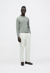 Férfi áll, világosszürke pulóvert, fehér nadrágot és sötétbarna mokaszint visel, egyszínű fehér háttér előtt.