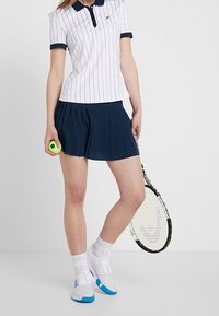 Vit och marinblå randig pikétröja, plisserad marinblå kjol, vita sportskor med blå detaljer, håller en grön tennisboll och racket.