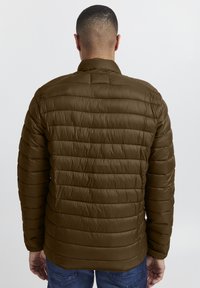 Olivengrøn dunjakke med quiltet tekstur, der har en ståkrave og elastiske manchetter, båret af en person med kort hår.