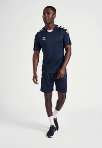 Tenue de sport bleu marine comprenant un maillot à manches courtes avec des accents blancs et un short assorti. Portée avec des chaussures noires et des chaussettes blanches.