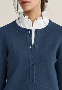 Kvinna som bär en vit blus med volangkrage under en knäppt marinblå kofta, med lösa hårstrån runt ansiktet.