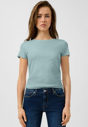 Jeune femme debout, face à l'avant, portant une chemise à manches courtes bleu clair et un jean bleu foncé sur un fond uni.