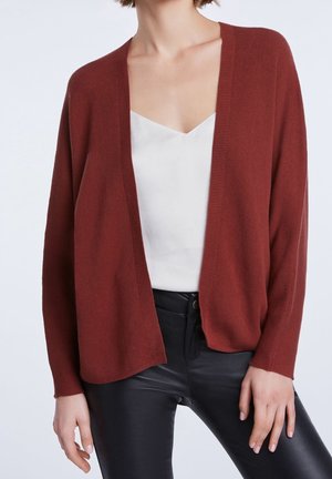 Frau trägt eine rostfarbene, offene Strickjacke, ein weißes V-Ausschnitt-Top und schwarze Lederhosen, stehend vor einem neutralen hellen Hintergrund.