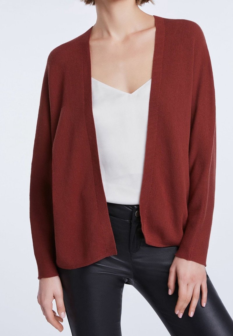 Femme portant un cardigan ouvert couleur rouille, un haut blanc à col en V et un pantalon en cuir noir, debout devant un fond clair uni.