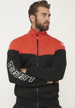 Muž s vousy, který má na sobě červeno-černou bundu na zip s nápisem "LEGEA" na levém rukávu, se zkříženýma rukama, dívá se stranou.