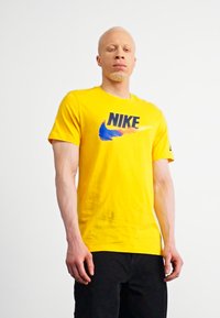 Geltona medvilninė marškinė su mėlynu ir juodu Nike logotipu, trumpomis rankovėmis ir klasikiniu apvaliu kaklu. Minimalistinis dizainas, be raštų.