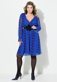 Robe bleue à motif avec un décolleté en V, des manches longues et une ceinture en fleur noire. Associée à des collants noirs et des escarpins bleus à bout pointu.