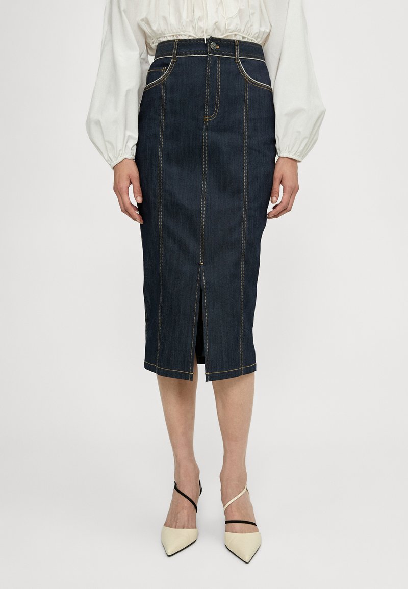 Jupe crayon en denim bleu foncé avec des coutures jaunes, fente frontale, taille haute et design ajusté. Portée avec des talons aiguilles de couleur claire.